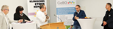 Fünf Personen stehen an Tischen und diskutieren über Ethik in der Medizin. Im Hintergrund stehen Roll ups zum Thema Ethik und der Klink.